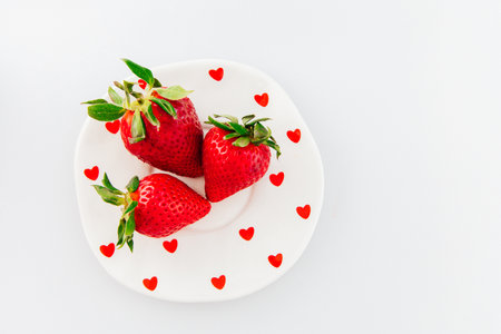 Bright red strawberries rest on a white plate adorned with playful heart shapes creating a romantic and inviting atmosphere perfect for sharing.の写真素材