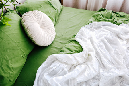 Soft green bed linens surround a white blanket and decorative pillow in a warm, inviting bedroom setting.の写真素材