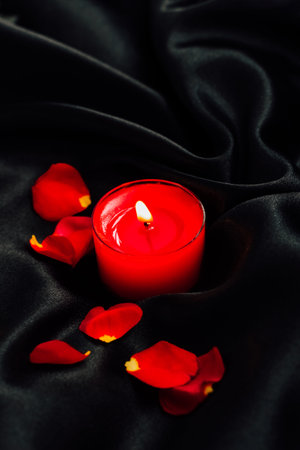 A gentle red candle flickers on a smooth black silk fabric with rose petals scattered around.の写真素材