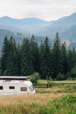 A vintage caravan rests in a green field surrounded by tall trees and mountains. The tranquil...
