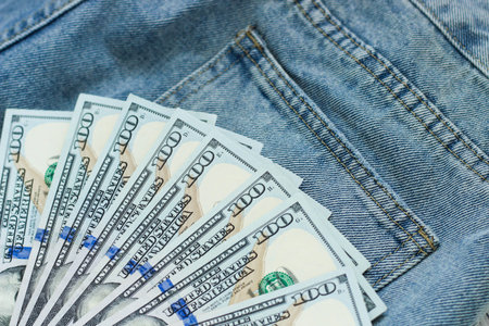 A collection of hundred dollar bills is arranged in a fan shape resting on blue denim fabric with a...
