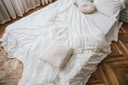 Soft white bedding is artfully arranged on a wooden floor, illuminated by natural light streaming through sheer curtains.の写真素材