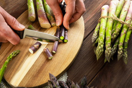 Hands skillfully chop asparagus spears on a wooden cutting board while more asparagus lies nearby in a kitchen.の写真素材
