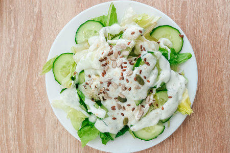 A vibrant salad featuring crunchy cucumbers and crisp lettuce topped with a creamy dressing and seeds. Enjoying a healthy meal on a wooden table.の写真素材