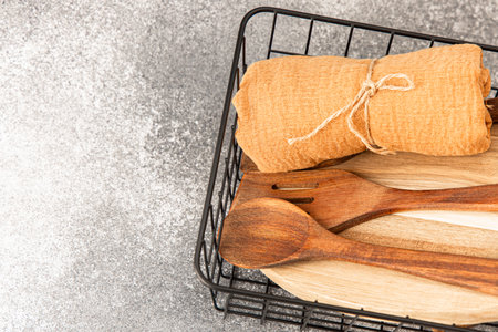 Wooden spoons and a folded textile are neatly placed in a wire basket on a gray background creating a rustic kitchen setting.の写真素材