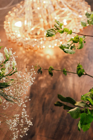 Floral arrangement featuring delicate white flowers and greenery illuminated by soft chandelier light in a charming space.の写真素材