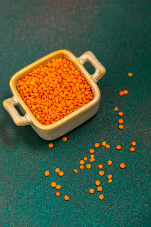 Small bowl filled with vibrant orange lentils placed on a green surface some lentils scattered around showcasing their texture and color.の写真素材