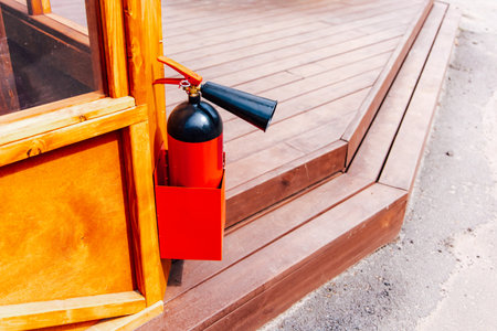A bright red fire extinguisher is securely mounted on a wooden post beside a wooden deck. The scene is well-lit by daytime sun.の写真素材