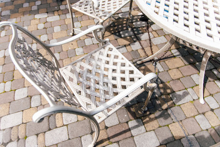 A metal chair with intricate designs and a matching table sit on a sunlit cobblestone patio inviting relaxation and enjoyment.の写真素材