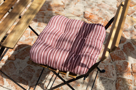 A striped cushion rests on a wooden chair casting a shadow on cobblestone in a bright outdoor space.の写真素材