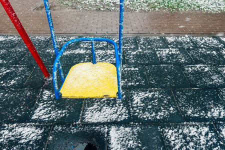 A vibrant yellow swing sits on a snowy playground floor while snowflakes gently fall around it creating a winter wonderland atmosphere.の写真素材