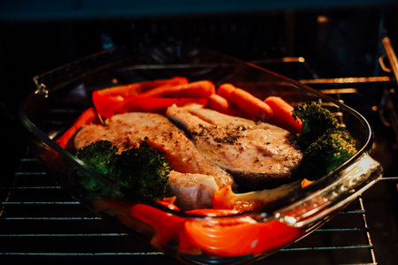 Salmon fillets seasoned and surrounded by vibrant carrots broccoli and bell peppers baking in the oven.の写真素材
