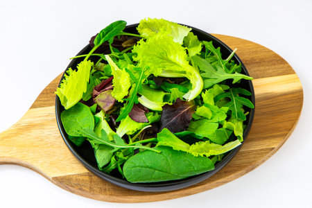 A vibrant assortment of leafy greens is arranged on a round black plate placed on a wooden serving board showcasing freshness and health.の写真素材