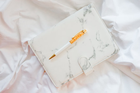 A stylish marble-patterned notebook lies open with a golden pen resting on its surface set against white fabric showcasing elegance and simplicity.の写真素材