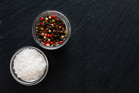 Various peppercorns and coarse salt are arranged in glass bowls atop a dark stone surface ready for cooking enhancements.の写真素材