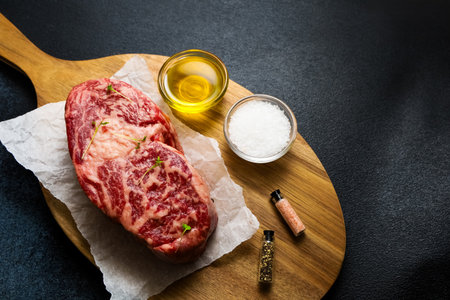 Two cuts of marbled steak are showcased on a wooden board alongside olive oil salt and spices for seasoning.の写真素材