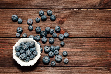 Fresh plump blueberries are scattered around a small white bowl on a rustic wooden table. The vibrant color contrasts beautifully with the wood.の写真素材