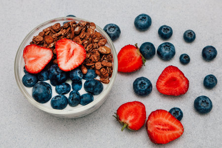 A bowl of yogurt topped with blueberries sliced strawberries and granola sits on a light gray surface surrounded by scattered berries.の写真素材