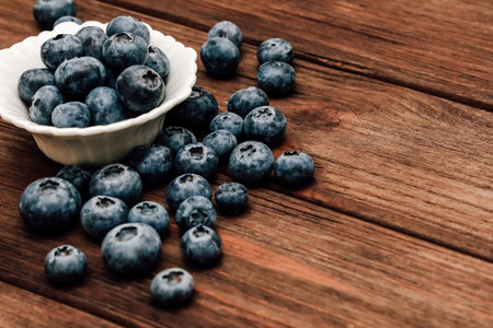 A bowl filled with fresh blueberries sits next to scattered berries on a weathered wooden table illuminated by soft morning light.の写真素材