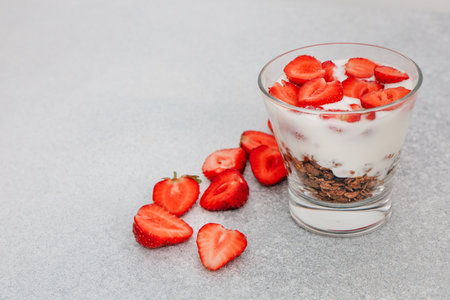 A yogurt parfait features layers of creamy yogurt crunchy granola and vibrant fresh strawberries placed on a light gray surface.の写真素材