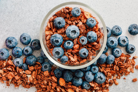 A glass bowl filled with crunchy granola topped with fresh blueberries is placed on a light surface creating an inviting breakfast option.の写真素材