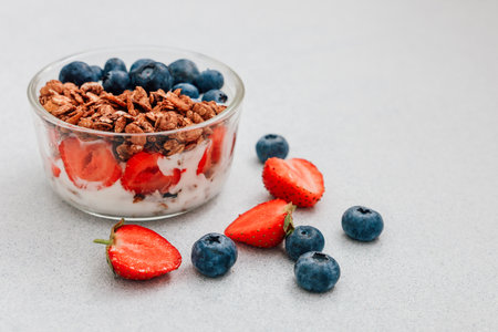 A tasty parfait is prepared with layers of yogurt fresh strawberries blueberries and crunchy granola on a light gray background.の写真素材