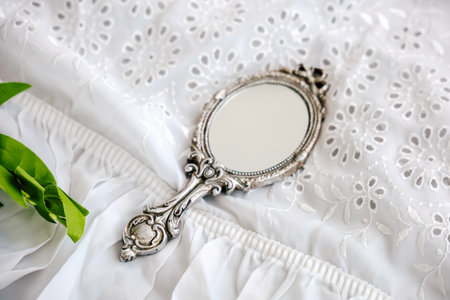 A decorative vintage hand mirror rests on a soft white embroidered fabric surrounded by delicate green leaves creating an elegant arrangement.の写真素材