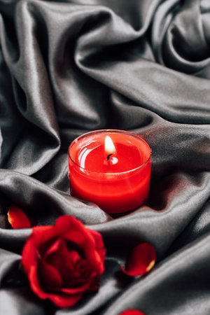 A bright red candle glows gently on smooth black satin fabric surrounded by soft rose petals and a deep red rose creating a romantic ambiance.の写真素材