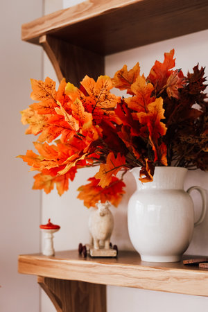 A white pitcher displays bright orange leaves on a wooden shelf capturing the essence of autumn. The warm colors create a cozy atmosphere.の写真素材
