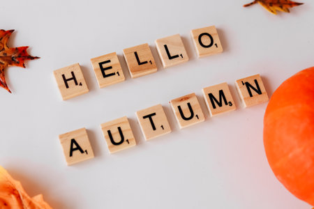 Wooden letter tiles spell out a warm greeting for autumn accompanied by colorful leaves and a pumpkin on a white background.の写真素材