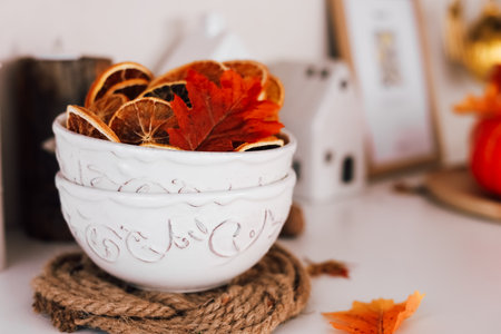 Two white decorative bowls are filled with dried orange slices and vibrant autumn leaves resting on a rustic table.の写真素材
