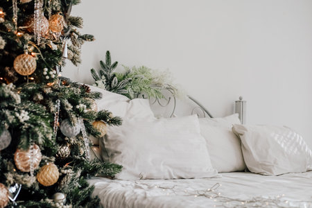 A beautifully decorated Christmas tree stands beside a cozy bed covered in white pillows and blankets creating a warm festive atmosphere.の写真素材