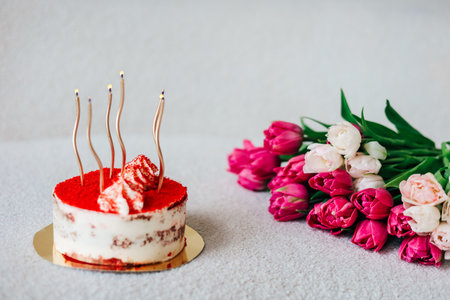 A round red velvet cake featuring four candles sits next to a bouquet of pink tulips creating a festive atmosphere for a special occasion.の写真素材