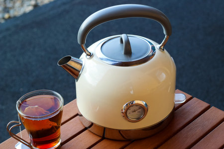 A modern kettle sits next to a transparent cup filled with dark tea on a wooden surface in a sunny outdoor setting.の写真素材