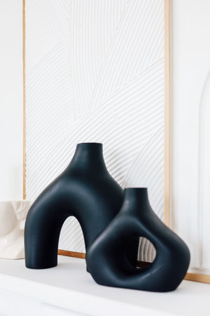 Stylish black vases are displayed on a clean shelf contrasting with textured wall art in a contemporary room.の写真素材