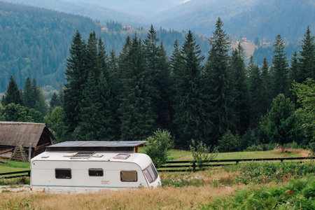 A caravan is stationed in a lush green meadow with towering trees and Carpathiansmountains in the backdrop.の写真素材