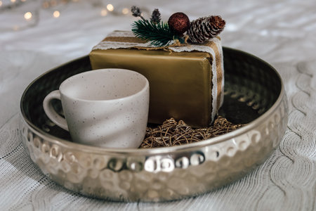A delicate cup rests beside a beautifully wrapped gift on a shiny tray amidst a warm and inviting atmosphere. Soft lights enhance the cozy feel.の写真素材