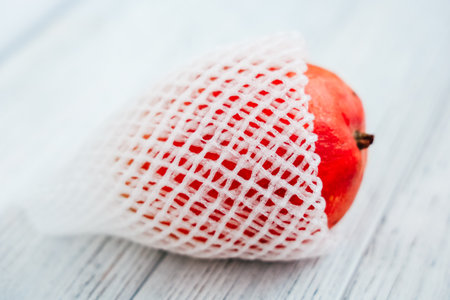 A vibrant red apple is carefully wrapped in white protective netting resting on a light wooden surface showcasing its fresh appearance.の写真素材
