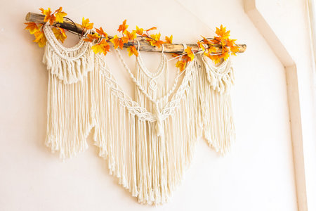 A beautiful macrame wall hanging with fringes and intricate patterns is adorned with vibrant autumn leaves creating a warm atmosphere indoors.の写真素材