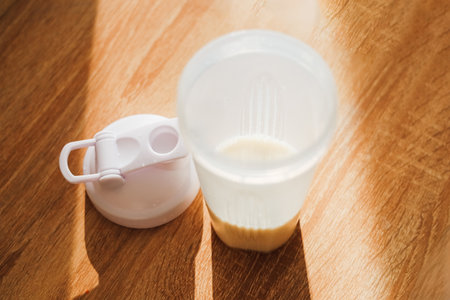 protein with a lid sits on a wooden table filled with a light colored smoothie in the morning sunlight.の写真素材