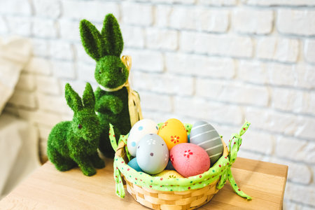 Brightly colored Easter eggs are arranged in a woven basket beside two green decorative bunnies made of moss on a wooden surface.の写真素材