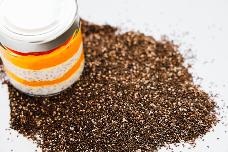 Layers of vibrant ingredients sit in a glass jar while chia seeds are scattered on a white surface nearby creating a colorful culinary display.の写真素材