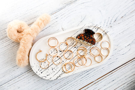 Gold rings and a hair clip are neatly arranged on a stylish tray next to a cute fuzzy accessory in a bright space.の写真素材