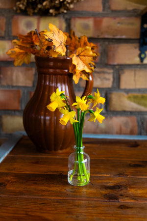 Colorful yellow flowers are arranged in a small glass vase sitting on a wooden table beside autumn leaves.の写真素材
