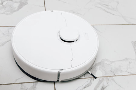 A white robot vacuum cleaner is actively cleaning a smooth tiled floor in a well lit room during the afternoon hours showcasing modern technology.の写真素材