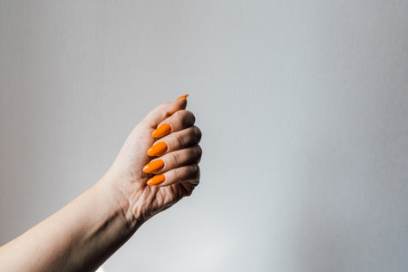 A hand displays long manicured orange nails clenched into a fist against a soft white background showcasing vibrant nail art.の写真素材