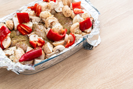 Fresh chicken is seasoned and topped with sliced red bell peppers and mushrooms arranged neatly in a foil tray on a wooden table.の写真素材