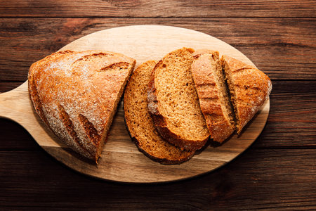 Warm freshly baked brown bread is displayed on a wooden cutting board with several slices cut and arranged neatly showcasing its texture and color.の写真素材