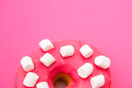 A vibrant donut topped with pink icing and small marshmallows rests on a bright pink background...