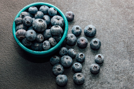A turquoise bowl filled with fresh blueberries rests on a textured surface with some berries...